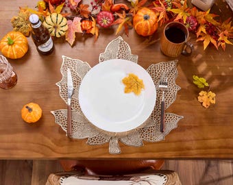 May include: A fall-themed table setting with a white plate, leaf-shaped placemat, and silverware. Decorative pumpkins, fall leaves, and a mug of coffee are arranged around the plate. A bottle of beer and a glass are also present.