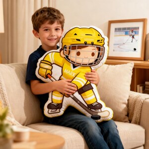 May include: A yellow and white hockey player pillow with the name "MARK" on the front. The pillow is held by a person sitting on a beige couch. A framed picture of a hockey player is in the background.