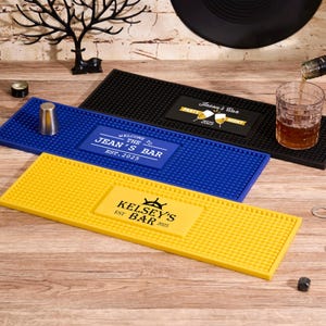 Puede incluir: Tres alfombrillas rectangulares para bar en negro, azul y amarillo, cada una con una superficie texturizada. Las alfombrillas presentan textos personalizados: "Jean's Bar", "Kelsey's Bar" y "Party Night". También se ve un vaso de chupito y una bebida que se vierte en un vaso.