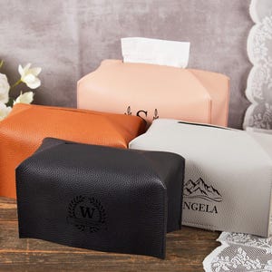 Może przedstawiać: Cztery skórzane osłony na pudełka z chusteczkami w różnych kolorach: czarnym, pomarańczowym, różowym i szarym. Każde pudełko ma prostokątny kształt z otworem u góry do dozowania chusteczek. Niektóre pudełka mają spersonalizowane wzory.