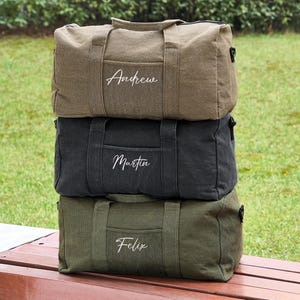 May include: Three canvas duffel bags stacked on top of each other. The top bag is brown with the name "Andrew" embroidered on it. The middle bag is black with the name "Martin" embroidered on it. The bottom bag is green with the name "Felix" embroidered on it.