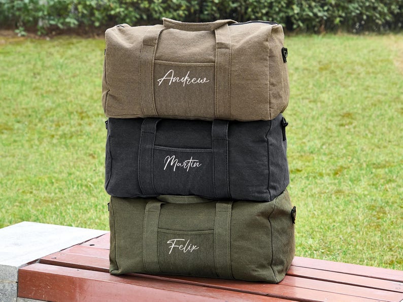 May include: Three canvas duffel bags stacked on top of each other. The top bag is brown with the name "Andrew" embroidered on it. The middle bag is black with the name "Martin" embroidered on it. The bottom bag is green with the name "Felix" embroidered on it.