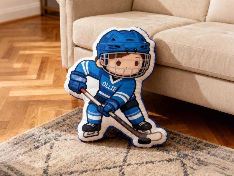 May include: A hockey-themed pillow in the shape of a cartoon hockey player, wearing a blue helmet and uniform. The player is holding a hockey stick and puck. The name "OLLIE" is printed on the jersey. The pillow is on a patterned rug.