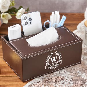 May include: A brown leatherette tissue box with compartments for a phone, charger, and pens. The box has white stitching and a personalized emblem with the letter "W" and the name "ROBERT".