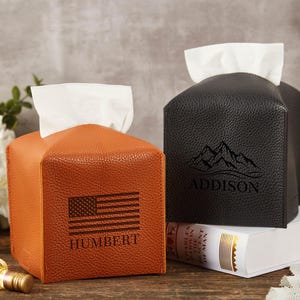 Puede incluir: Dos cajas de pañuelos de cuero, una naranja y otra negra, cada una con forma cuadrada y una abertura superior para los pañuelos. La caja naranja presenta una bandera estadounidense y el nombre "HUMBERT". La caja negra tiene un diseño de montaña y el nombre "ADDISON".
