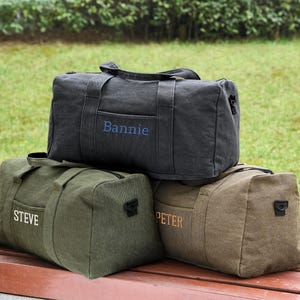 May include: Three canvas duffel bags with personalized names. The top bag is black with the name "Bannie" embroidered in blue. The middle bag is green with the name "Steve" embroidered in white. The bottom bag is brown with the name "Peter" embroidered in orange.