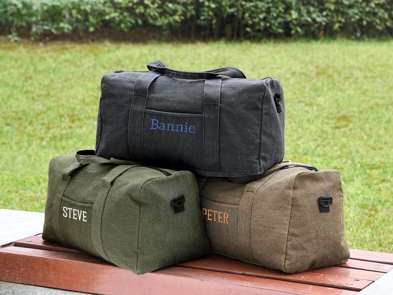 May include: Three canvas duffel bags with personalized names. The top bag is black with the name "Bannie" embroidered in blue. The middle bag is green with the name "Steve" embroidered in white. The bottom bag is brown with the name "Peter" embroidered in orange.