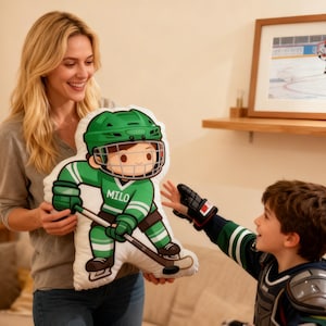 May include: A green and white hockey player pillow with the name "Milo" on the jersey. The pillow is held by a person, and a child reaches for it. A framed picture of a hockey game is on the wall.