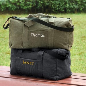 May include: Two canvas duffel bags stacked on top of each other. The top bag is green and has the name "Thomas" embroidered on it. The bottom bag is black and has the name "Janet" embroidered on it.