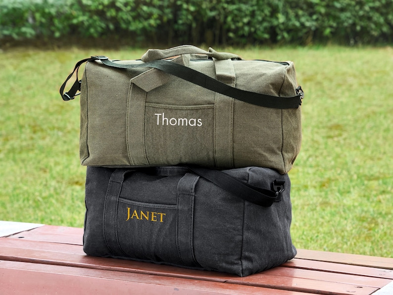 May include: Two canvas duffel bags stacked on top of each other. The top bag is green and has the name "Thomas" embroidered on it. The bottom bag is black and has the name "Janet" embroidered on it.