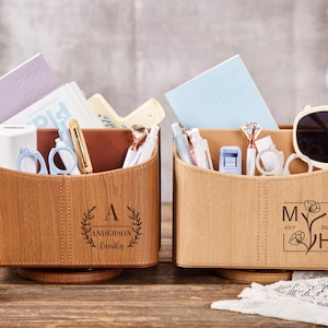 Puede incluir: Dos organizadores de escritorio de madera con varios artículos de papelería. Uno tiene el texto "A Anderson Family" dentro de una corona de laurel. El otro tiene el texto "MH July 2025" con un diseño floral. Ambos están sobre una base giratoria.