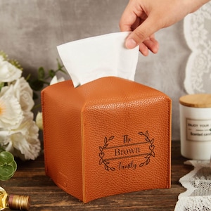 Può includere: Un porta fazzoletti quadrato e strutturato, di colore arancione, con un motivo floreale decorativo e la scritta "The Brown Family". I fazzoletti bianchi sporgono dalla parte superiore. Una mano sta tirando un fazzoletto.
