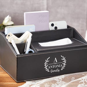 Puede incluir: Organizador de escritorio negro con costuras blancas, con bolígrafos, un teléfono y un bloc de notas. El organizador tiene una caja de pañuelos y está personalizado con "A - The Antonia Family" en un diseño de corona blanca.