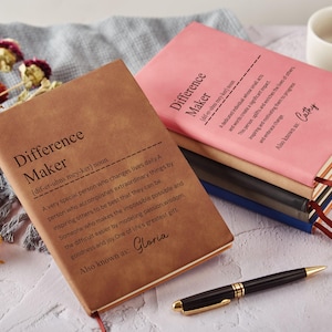 Puede incluir: Dos cuadernos de tapa dura con el texto "Difference Maker" en las cubiertas. Uno es marrón, el otro rosa. Un bolígrafo negro y dorado descansa cerca de los cuadernos. Flores secas y una taza blanca en el fondo.
