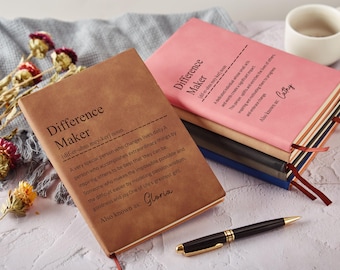 May include: Two hardcover notebooks with the text "Difference Maker" on the covers. One is brown, the other pink. A black and gold pen rests near the notebooks. Dried flowers and a white mug are in the background.