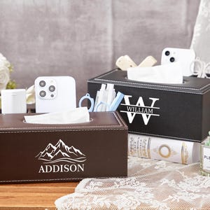 May include: Two rectangular tissue boxes are shown, one brown and one black. The brown box has a mountain graphic and the name "Addison." The black box features the name "William." Both boxes are filled with white tissues.