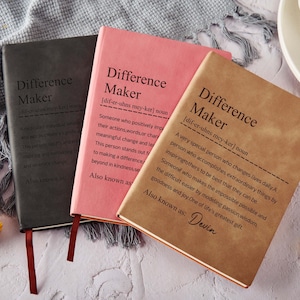 Puede incluir: Tres cuadernos de tapa dura en negro, rosa y marrón, cada uno con el texto "Difference Maker" y una definición. También se ven un bolígrafo negro y granos de café. Los cuadernos tienen cintas de marcador adjuntas.