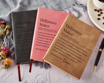 Può includere: Tre quaderni con copertina rigida neri, rosa e marroni, ciascuno con il testo "Difference Maker" e una definizione. Sono visibili anche una penna nera e chicchi di caffè. I quaderni hanno segnalibri a nastro attaccati.