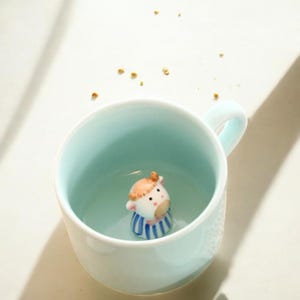 以下が含まれることがあります： 薄い青色のセラミック製のマグカップの中に白い牛の置物が入っています。牛は茶色の目、茶色の鼻、茶色の斑点があります。マグカップは白い縁と白い取っ手があります。