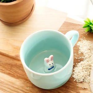 以下が含まれることがあります： 薄い青色のセラミック製のマグカップの中に、白いウサギの置物が入っています。ウサギはピンクの耳と青い蝶ネクタイをしています。