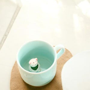 以下が含まれることがあります： 薄い青色のセラミック製のマグカップの中に、小さな白いセラミック製の動物の置物が置かれています。置物は緑色のスカーフをしています。