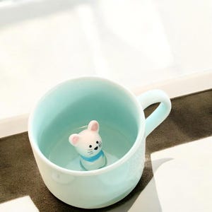 以下が含まれることがあります： 薄い青色のセラミック製のマグカップの中に、白いセラミック製のネズミの置物が入っています。ネズミはピンクの鼻と青いマフラーをしています。
