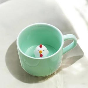 以下が含まれることがあります： 薄い緑色のセラミック製のマグカップの中に、小さな白い鶏の置物が入っています。鶏は赤い鶏冠とくちばしをしています。