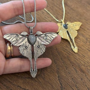 May include: A silver-colored moth pendant necklace with a teardrop-shaped stone in the center. The moth's wings are detailed with intricate patterns. A gold-colored moth pendant necklace is also visible.