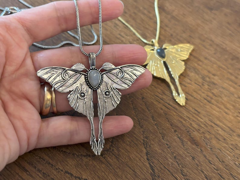 May include: A silver-colored moth pendant necklace with a teardrop-shaped stone in the center. The moth's wings are detailed with intricate patterns. A gold-colored moth pendant necklace is also visible.