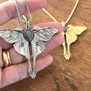 May include: Silver-toned moth pendant necklace with a gemstone center, held in a hand. The pendant has detailed wing patterns and a long, stylized body. A gold-toned moth pendant necklace is in the background.