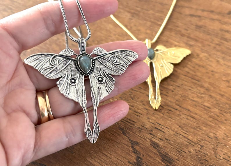 May include: Silver-toned moth pendant necklace with a gemstone center, held in a hand. The pendant has detailed wing patterns and a long, stylized body. A gold-toned moth pendant necklace is in the background.