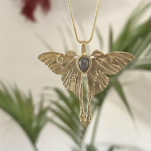 May include: A gold-colored metal pendant necklace featuring a detailed moth design. The moth has outstretched wings and a central teardrop-shaped gray stone. The necklace chain is also gold-colored, and the background is blurred with green foliage.