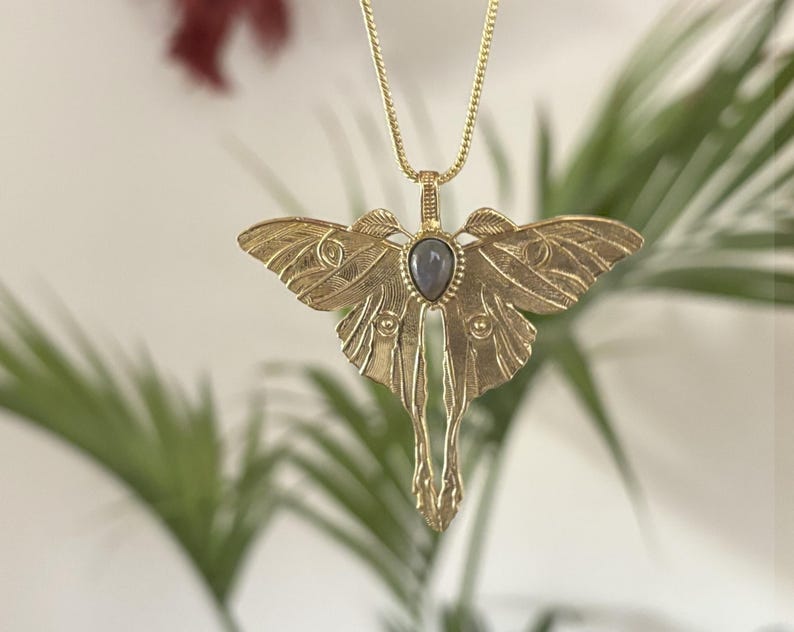 May include: A gold-colored metal pendant necklace featuring a detailed moth design. The moth has outstretched wings and a central teardrop-shaped gray stone. The necklace chain is also gold-colored, and the background is blurred with green foliage.