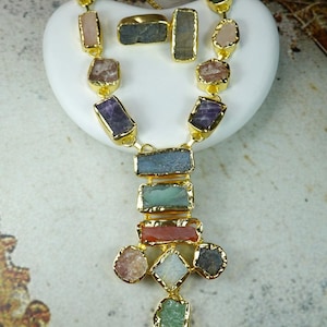 Puede incluir: Un conjunto de collar y anillo dorados con piedras preciosas de colores de forma irregular. El collar tiene un colgante con un grupo de piedras. El anillo tiene una piedra rectangular. Las piedras son de color púrpura, verde, rosa y azul.