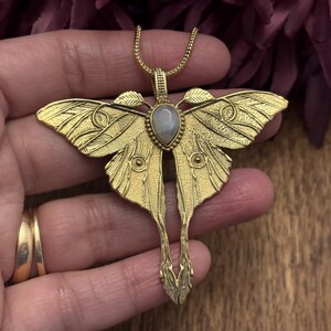 May include: A gold-colored moth pendant necklace with a detailed wing design and a teardrop-shaped, light-colored stone in the center. The pendant is on a delicate chain.
