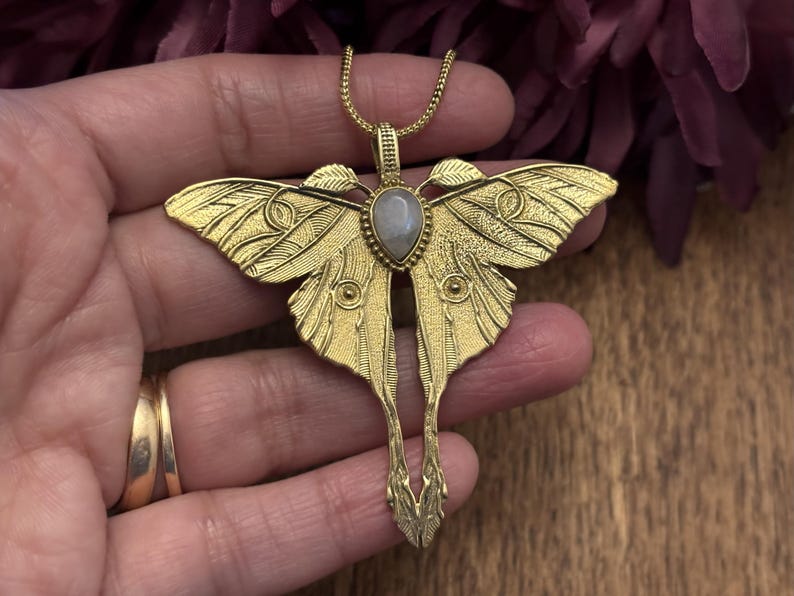 May include: A gold-colored moth pendant necklace with a detailed wing design and a teardrop-shaped, light-colored stone in the center. The pendant is on a delicate chain.