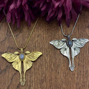 May include: Two moth-shaped pendant necklaces. One is gold-toned, the other silver-toned. Each pendant has a central gemstone and hangs from a delicate chain. The pendants are displayed on a wooden surface, with dark purple flowers in the background.