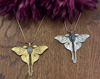Puede incluir: Dos collares con colgantes en forma de polilla. Uno es dorado, el otro plateado. Cada colgante tiene una piedra preciosa central y cuelga de una delicada cadena. Los colgantes se exhiben sobre una superficie de madera, con flores moradas oscuras en el fondo.
