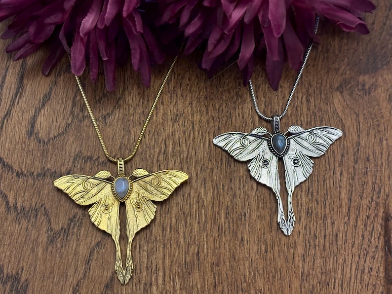 May include: Two moth-shaped pendant necklaces. One is gold-toned, the other silver-toned. Each pendant has a central gemstone and hangs from a delicate chain. The pendants are displayed on a wooden surface, with dark purple flowers in the background.