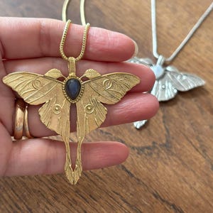 May include: A gold-colored moth pendant necklace with a dark blue gemstone center. The pendant has detailed wing and body designs. A silver-colored moth pendant necklace is in the background.