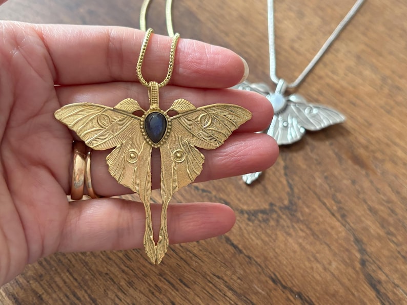 May include: A gold-colored moth pendant necklace with a dark blue gemstone center. The pendant has detailed wing and body designs. A silver-colored moth pendant necklace is in the background.