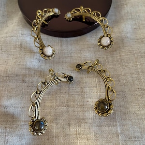 Könnte beinhalten: Vier antike, goldfarbene Ohrklemmen mit dekorativen Filigranarbeiten und Edelsteinakzenten. Zwei Manschetten haben weiße Edelsteine, und zwei haben dunkelbraune Edelsteine. Die Ohrklemmen sind auf einer neutralen Leinenoberfläche angeordnet.