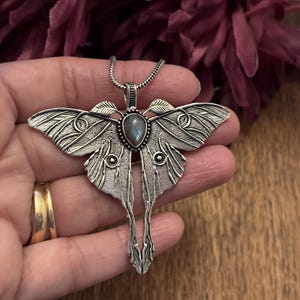 May include: Silver-toned moth pendant necklace. The pendant features intricate wing detailing, a central gemstone, and a delicate chain. The moth design is a detailed representation of the insect, with a focus on the wings and body.