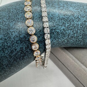 May include: Two bracelets are shown. One has clear, round gemstones in gold settings, the other has clear, square-cut gemstones in a silver metal setting. Both are displayed on a textured blue surface.