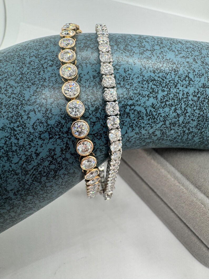 May include: Two bracelets are shown. One has clear, round gemstones in gold settings, the other has clear, square-cut gemstones in a silver metal setting. Both are displayed on a textured blue surface.