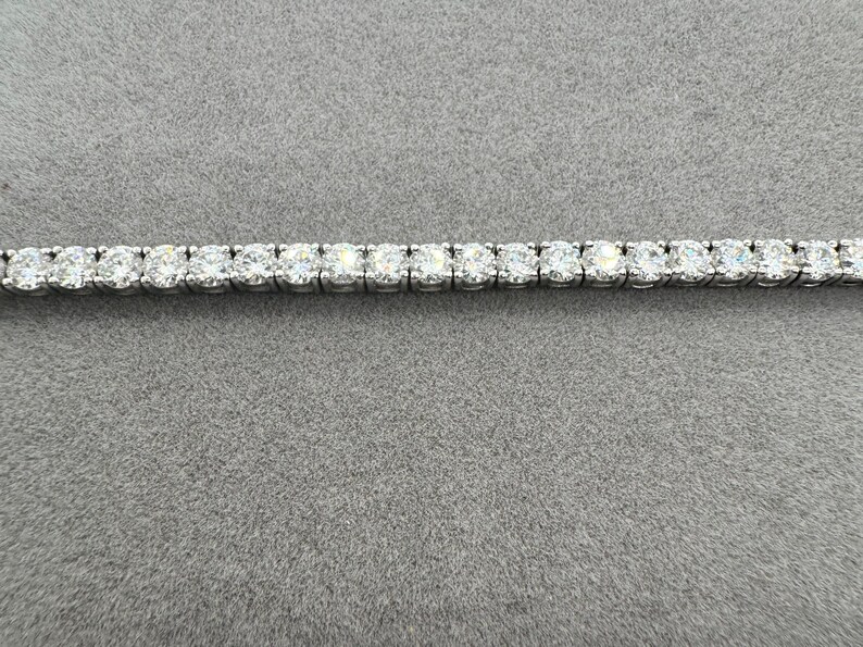 May include: A tennis bracelet with a continuous row of round-cut diamonds set in a silver-coloured metal. The bracelet is shown against a grey background, highlighting the sparkle of the gemstones.