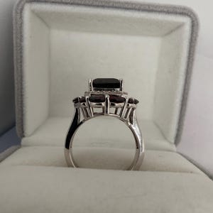 May include: A silver ring featuring a large, dark rectangular gemstone at its centre, encircled by smaller, round, dark red stones. The ring is presented in a grey velvet box.
