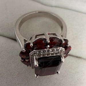 May include: A silver ring with a large, dark red rectangular gemstone at its centre, encircled by smaller, oval red gemstones. The ring has a detailed setting with small, clear accent stones.