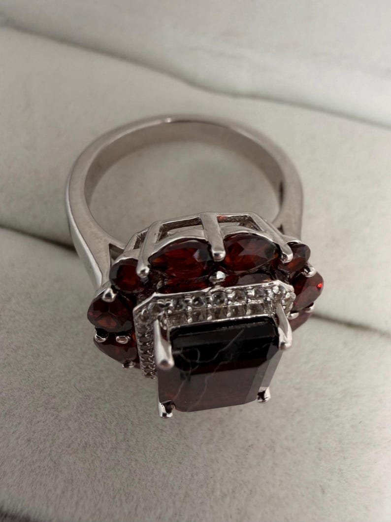 May include: A silver ring with a large, dark red rectangular gemstone at its centre, encircled by smaller, oval red gemstones. The ring has a detailed setting with small, clear accent stones.