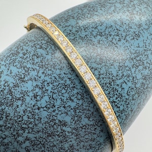 May include: A gold bangle bracelet with a row of clear gemstones. The bracelet is set against a textured, blue background. The gemstones are evenly spaced along the top of the bracelet.